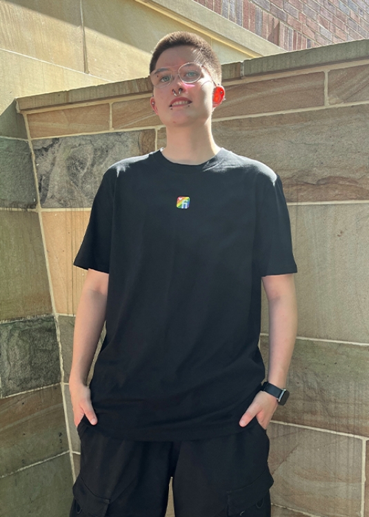 Model vor Steinwand mit schwarzem T-Shirt mit Finanz Informatik Logo in Regenbodenfarben